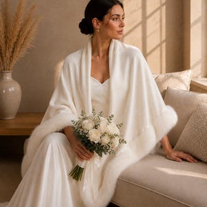 Bride sitting on a sofa wearing an ivory faux fur bridal shawl, soft wedding wrap styled with a modern bridal gown and white bouquet.