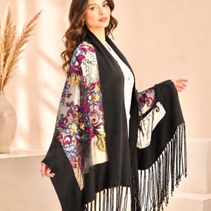 A black shawl with a floral patterned sheer panel and fringe trim. The sheer panel features a colorful floral design with yellow, purple, and blue flowers. The shawl is draped over a white outfit.