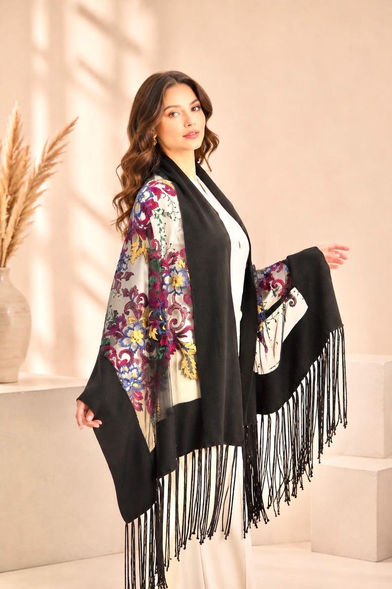 A black shawl with a floral patterned sheer panel and fringe trim. The sheer panel features a colorful floral design with yellow, purple, and blue flowers. The shawl is draped over a white outfit.