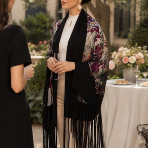 A woman wearing a black shawl with fringe and floral patterned sleeves. The shawl is draped over a white top and beige pants. The background includes a table with flowers and two other people.