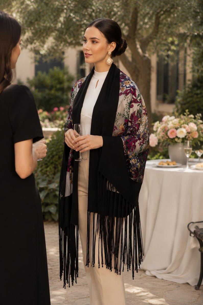 A woman wearing a black shawl with fringe and floral patterned sleeves. The shawl is draped over a white top and beige pants. The background includes a table with flowers and two other people.