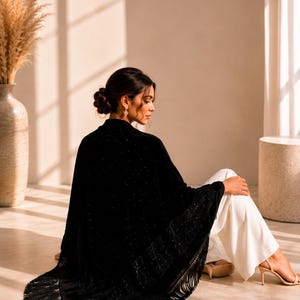 Woman wearing a luxury black velvet shawl adorned with crystal embellishments and long fringe, elegant wedding guest wrap styled in a bright editorial interior.