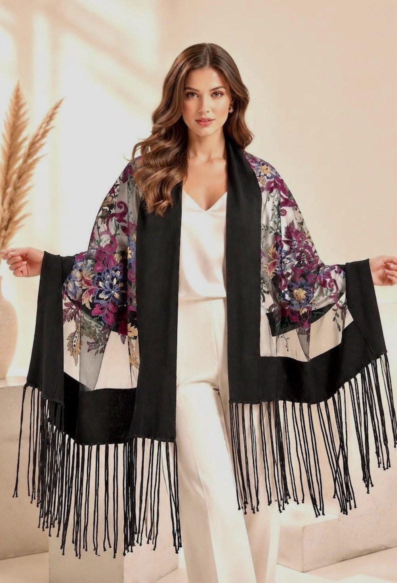 Luxury embroidered tulle shawl with black wool knit edges and long fringe details, featuring floral embroidery in burgundy, gold and blue tones. Elegant evening wrap styled for weddings, formal events and sophisticated occasion wear in a soft editorial studio setting.