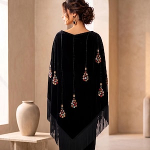 A black velvet poncho featuring a V-shaped hemline with long black fringe trim. The garment is adorned with vertical beaded strands that end in teardrop-shaped pendants encrusted with multicolored crystals and small metallic accents.