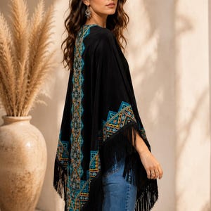 Black shawl with a geometric pattern in teal, orange, and gold. The poncho has long black fringe along the bottom and sleeves. The model is wearing blue jeans.
