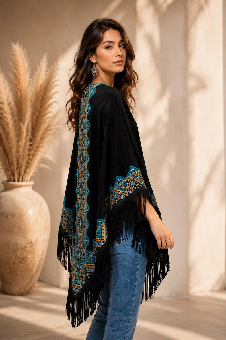 Black shawl with a geometric pattern in teal, orange, and gold. The poncho has long black fringe along the bottom and sleeves. The model is wearing blue jeans.