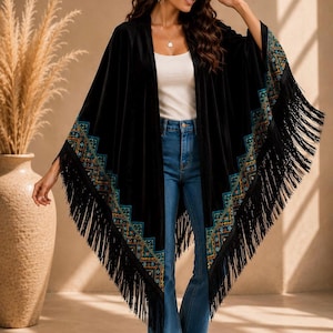 A black velvet shawl style wrap with colorful embroidered trim and long fringe. The wrap is worn over a white top and blue jeans. The sleeves are wide and flowing, and the trim features geometric patterns.