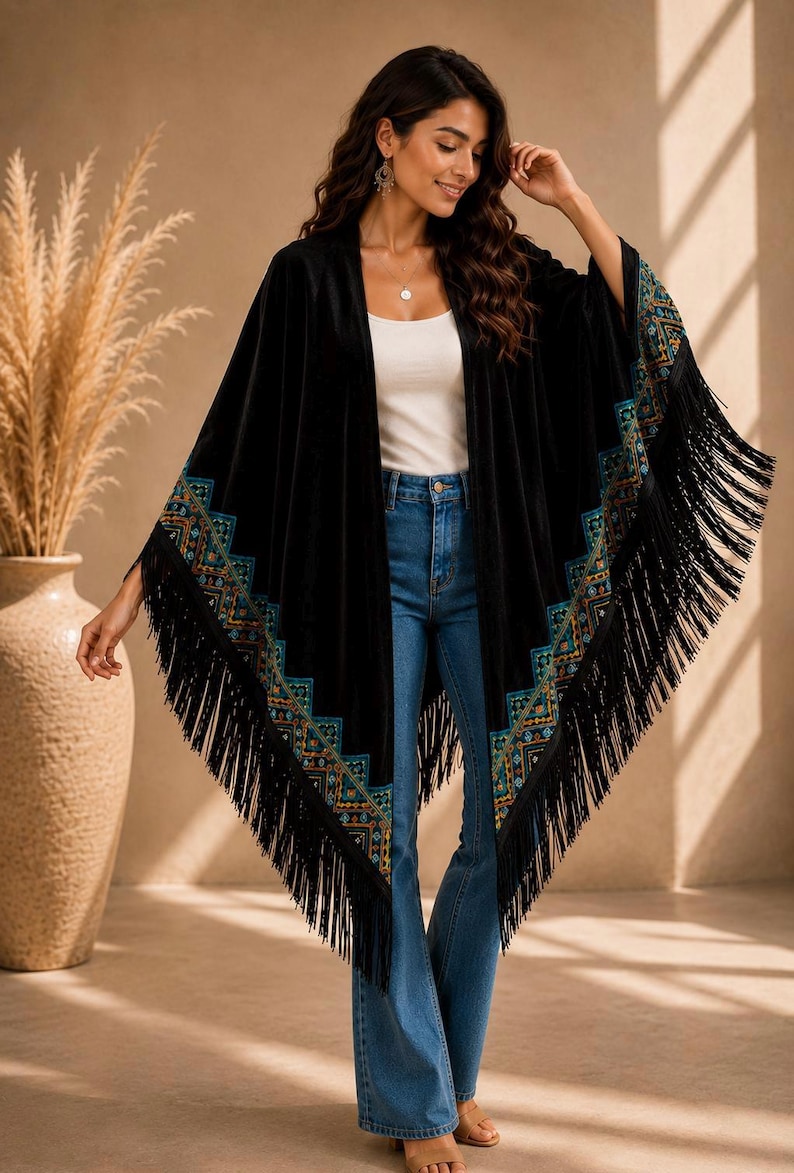 A black velvet shawl style wrap with colorful embroidered trim and long fringe. The wrap is worn over a white top and blue jeans. The sleeves are wide and flowing, and the trim features geometric patterns.