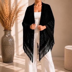 A black velvet wrap featuring intricate rhinestone embellishments in a geometric pattern and long fringe trim along the hem. The garment is worn over a white sleeveless top and matching wide-leg trousers.