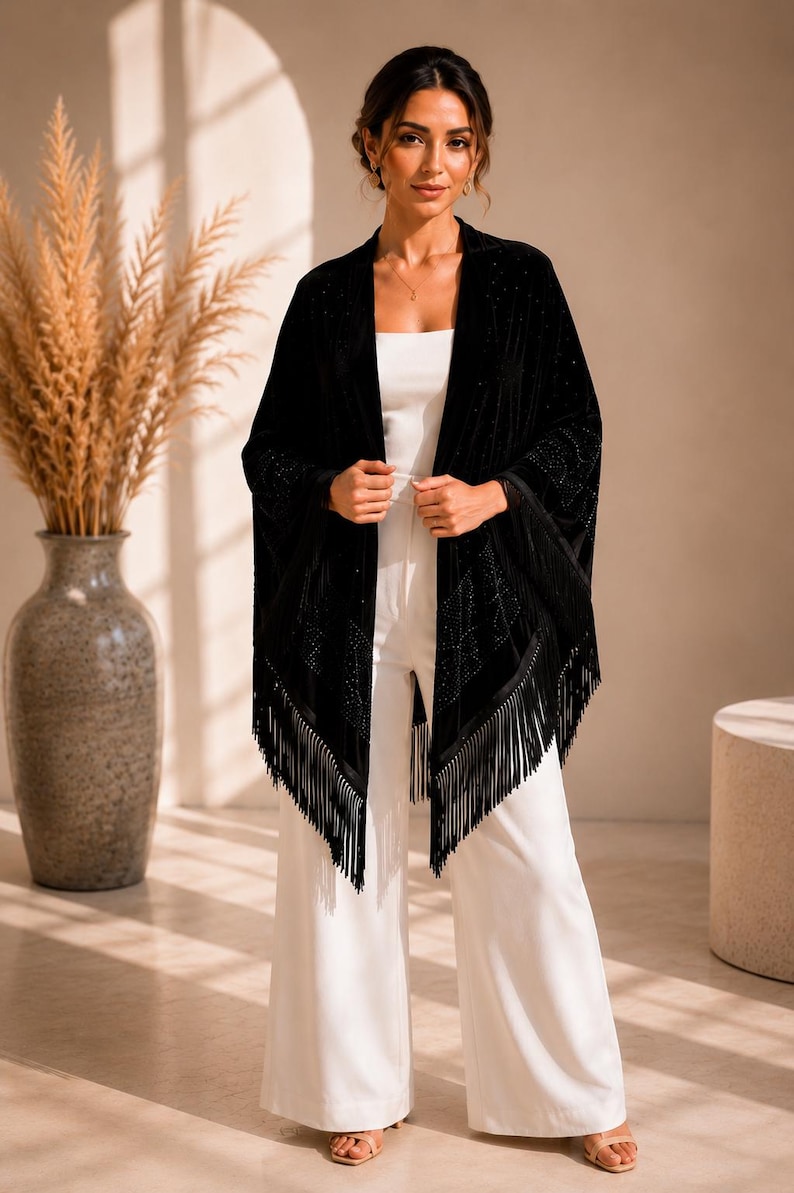A black velvet wrap featuring intricate rhinestone embellishments in a geometric pattern and long fringe trim along the hem. The garment is worn over a white sleeveless top and matching wide-leg trousers.