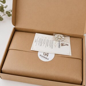 An open cardboard box containing a wrapped package, a decorative brooch, and a card. The package is wrapped in brown paper. A white sticker with the word "MANDORLE" is on the package. The brooch is silver and has a floral design.