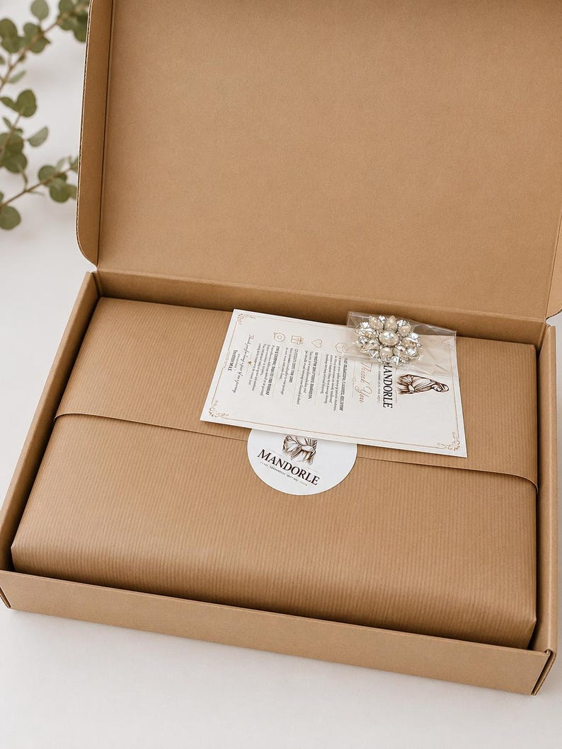An open cardboard box containing a wrapped package, a decorative brooch, and a card. The package is wrapped in brown paper. A white sticker with the word "MANDORLE" is on the package. The brooch is silver and has a floral design.