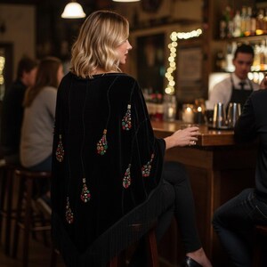 Black velvet shawl with colorful beaded embellishments in a teardrop design. The poncho has a fringed hem and is worn in a bar setting. The design features a series of beaded teardrops.