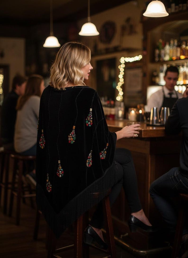 Black velvet shawl with colorful beaded embellishments in a teardrop design. The poncho has a fringed hem and is worn in a bar setting. The design features a series of beaded teardrops.
