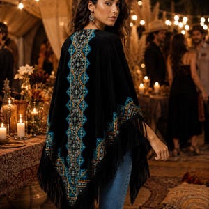 Black velvet shawl with intricate turquoise and orange geometric embroidery. The poncho features long fringe along the bottom edge. The garment is worn over blue jeans.