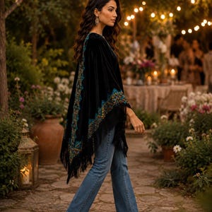 A black velvet shawl with intricate teal and gold embroidery and fringe trim. The poncho is paired with blue jeans and tan heels. The background features a garden setting with string lights and floral arrangements.