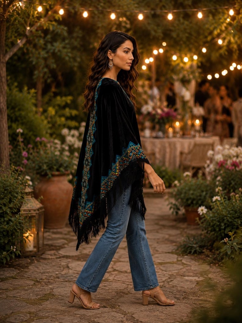 A black velvet shawl with intricate teal and gold embroidery and fringe trim. The poncho is paired with blue jeans and tan heels. The background features a garden setting with string lights and floral arrangements.