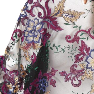 Close-up of a sheer garment with intricate floral embroidery. The design features burgundy, gold, blue, and green thread on a white background. A black collar is visible.