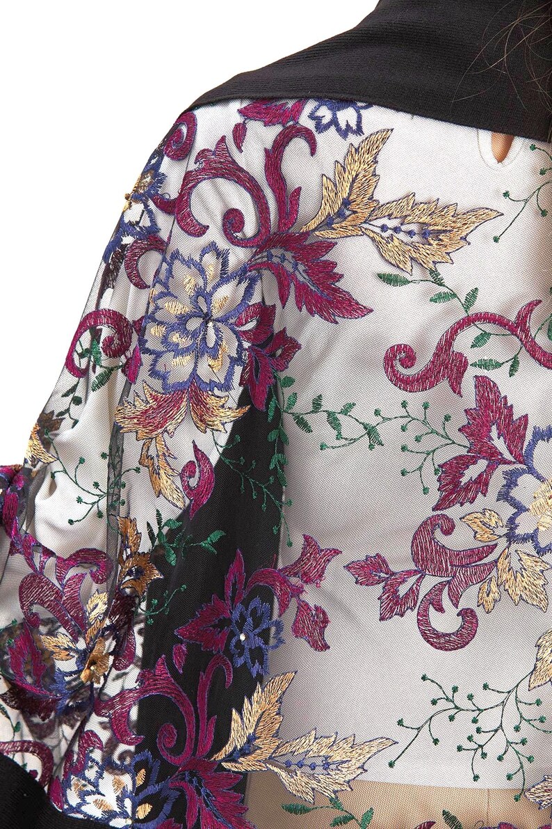 Close-up of a sheer garment with intricate floral embroidery. The design features burgundy, gold, blue, and green thread on a white background. A black collar is visible.