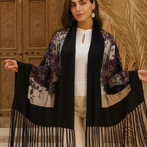 A black shawl with a floral pattern and fringe. The shawl has a black border and a sheer floral design in shades of purple, beige, and green. The shawl is worn over a white top and beige pants.