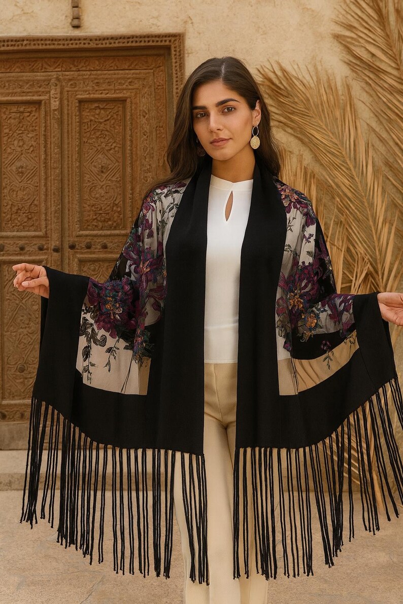 A black shawl with a floral pattern and fringe. The shawl has a black border and a sheer floral design in shades of purple, beige, and green. The shawl is worn over a white top and beige pants.