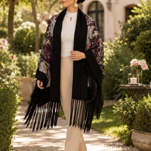 A woman wearing a floral print shawl with a black fringe scarf. The kimono features a sheer fabric with burgundy and purple floral patterns. The scarf is black and has long fringe. The woman is wearing a white top and beige pants.
