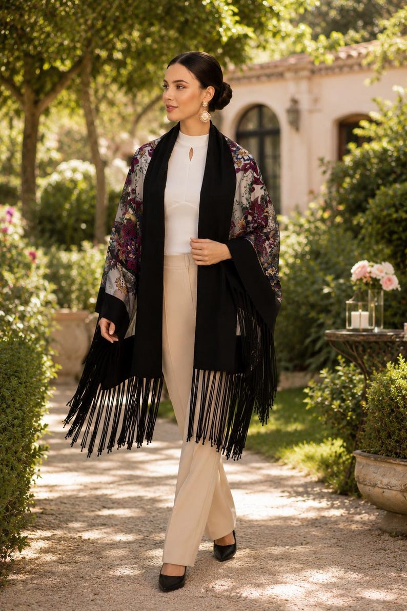 A woman wearing a floral print shawl with a black fringe scarf. The kimono features a sheer fabric with burgundy and purple floral patterns. The scarf is black and has long fringe. The woman is wearing a white top and beige pants.