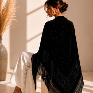 Elegant woman seated on a stone pedestal wearing a premium velvet shawl with crystal embellishments, sophisticated evening accessory with fringe edging.