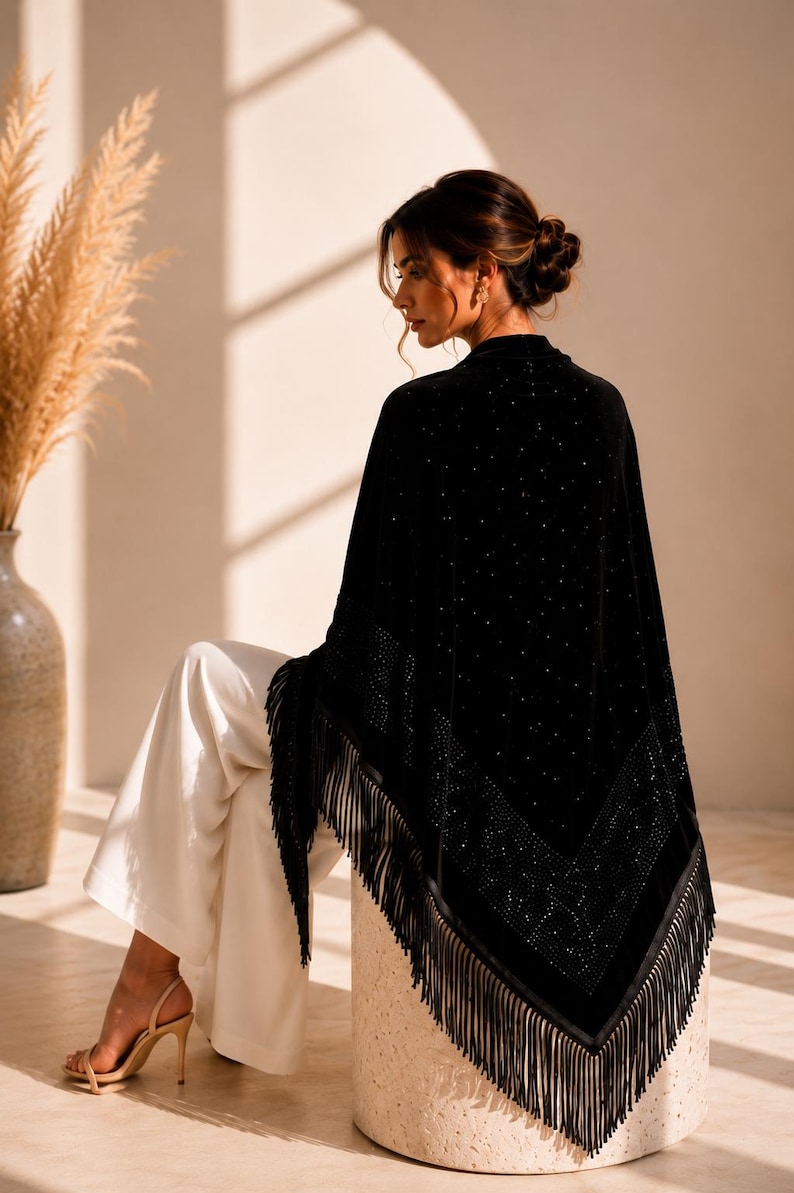 Elegant woman seated on a stone pedestal wearing a premium velvet shawl with crystal embellishments, sophisticated evening accessory with fringe edging.
