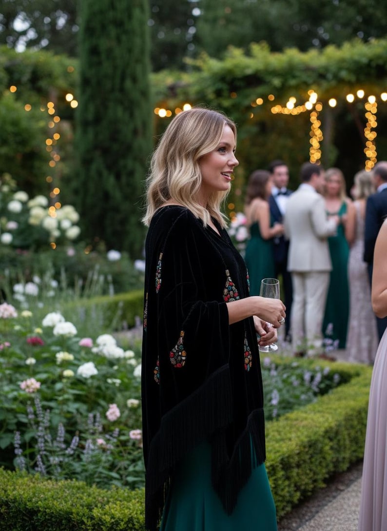 May include: A black velvet poncho featuring colorful embroidered paisley patterns and long fringe trim along the hemline, worn over a solid dark green floor-length dress. The garment has a loose, draped silhouette with wide sleeves.