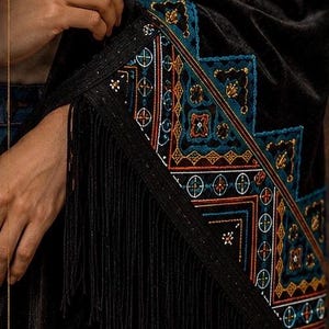 Black velvet shawl with intricate embroidered detailing in teal, orange, and white. The shawl features a geometric pattern along the border and is finished with a long fringe. The design is rich and detailed.