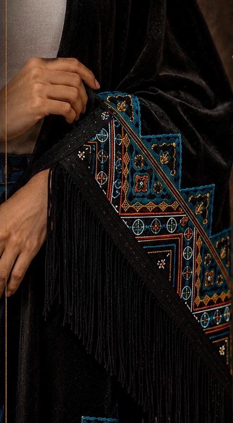 Black velvet shawl with intricate embroidered detailing in teal, orange, and white. The shawl features a geometric pattern along the border and is finished with a long fringe. The design is rich and detailed.