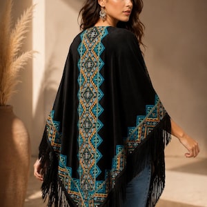 Black shawl with a geometric pattern in teal, orange, and gold. The poncho has a fringe trim along the bottom edge. The model is wearing blue jeans.