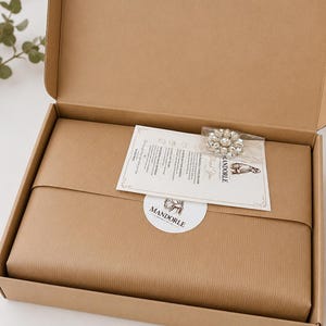 May include: An open brown cardboard shipping box containing a wrapped package secured with a round white sticker labeled MANDORLE. A rectangular thank you card and a small clear plastic bag with a crystal brooch are placed on top.