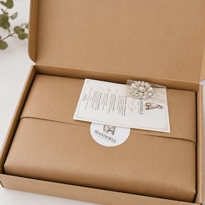 A brown cardboard box containing a wrapped package, a small card, and a brooch. The package is wrapped in brown paper and sealed with a white sticker that reads "MANDORLE". The brooch is silver-toned with clear crystals.