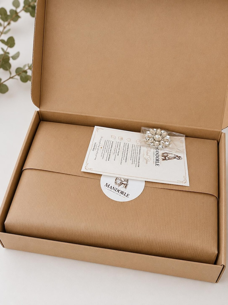 A brown cardboard box containing a wrapped package, a small card, and a brooch. The package is wrapped in brown paper and sealed with a white sticker that reads "MANDORLE". The brooch is silver-toned with clear crystals.