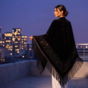 A black velvet shawl featuring intricate gold-toned embroidery and sparkling embellishments along the border. The triangular garment is finished with long, flowing black fringe trim along the edges, worn over simple white trousers.
