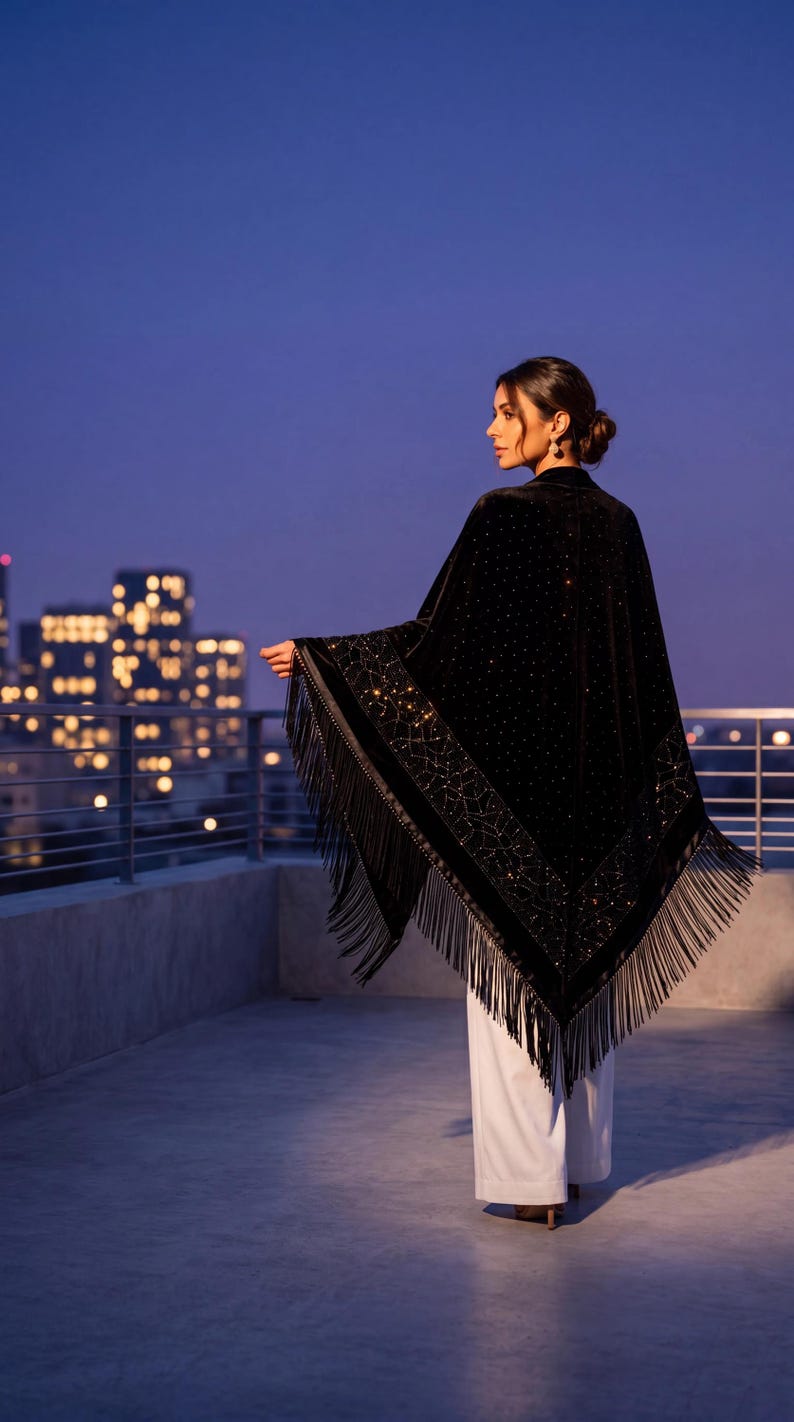 A black velvet shawl featuring intricate gold-toned embroidery and sparkling embellishments along the border. The triangular garment is finished with long, flowing black fringe trim along the edges, worn over simple white trousers.