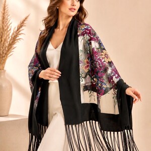 A black shawl with a floral design in shades of purple, yellow, and blue. The shawl has a fringe trim and is draped over the shoulders. The floral pattern is on a sheer panel.