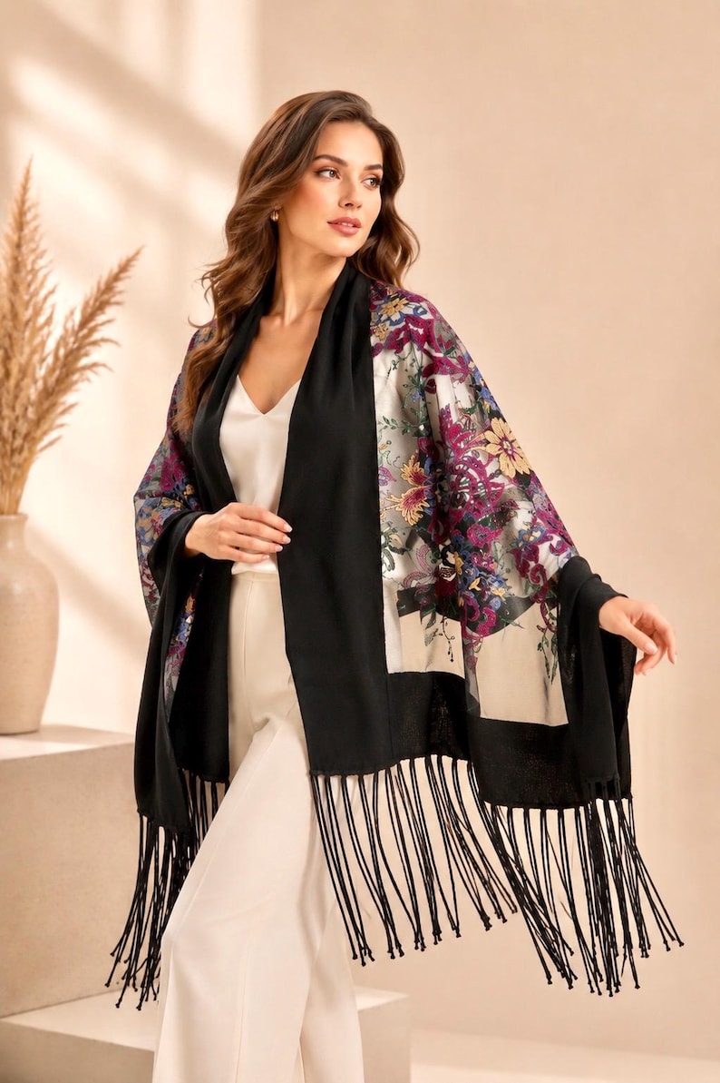 A black shawl with a floral design in shades of purple, yellow, and blue. The shawl has a fringe trim and is draped over the shoulders. The floral pattern is on a sheer panel.