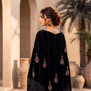 May include: A black velvet poncho featuring long fringe trim along the hem and vertical rows of dangling, teardrop-shaped embellishments adorned with multicolored crystals. The garment has a V-shaped silhouette and a smooth, dark fabric texture.