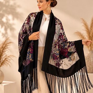 A floral print shawl with black trim and fringe. The shawl features a colorful floral pattern with burgundy, purple, and blue hues on a cream background. The black trim and fringe add a contrasting element to the design.