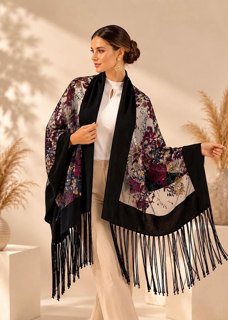 A floral print shawl with black trim and fringe. The shawl features a colorful floral pattern with burgundy, purple, and blue hues on a cream background. The black trim and fringe add a contrasting element to the design.