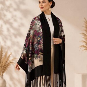 A floral embroidery shawl with black borders and fringe. The shawl features a colorful floral pattern on a light background. The black borders frame the floral design, and the fringe adds a decorative element. This accessory can be worn over the shoulders.