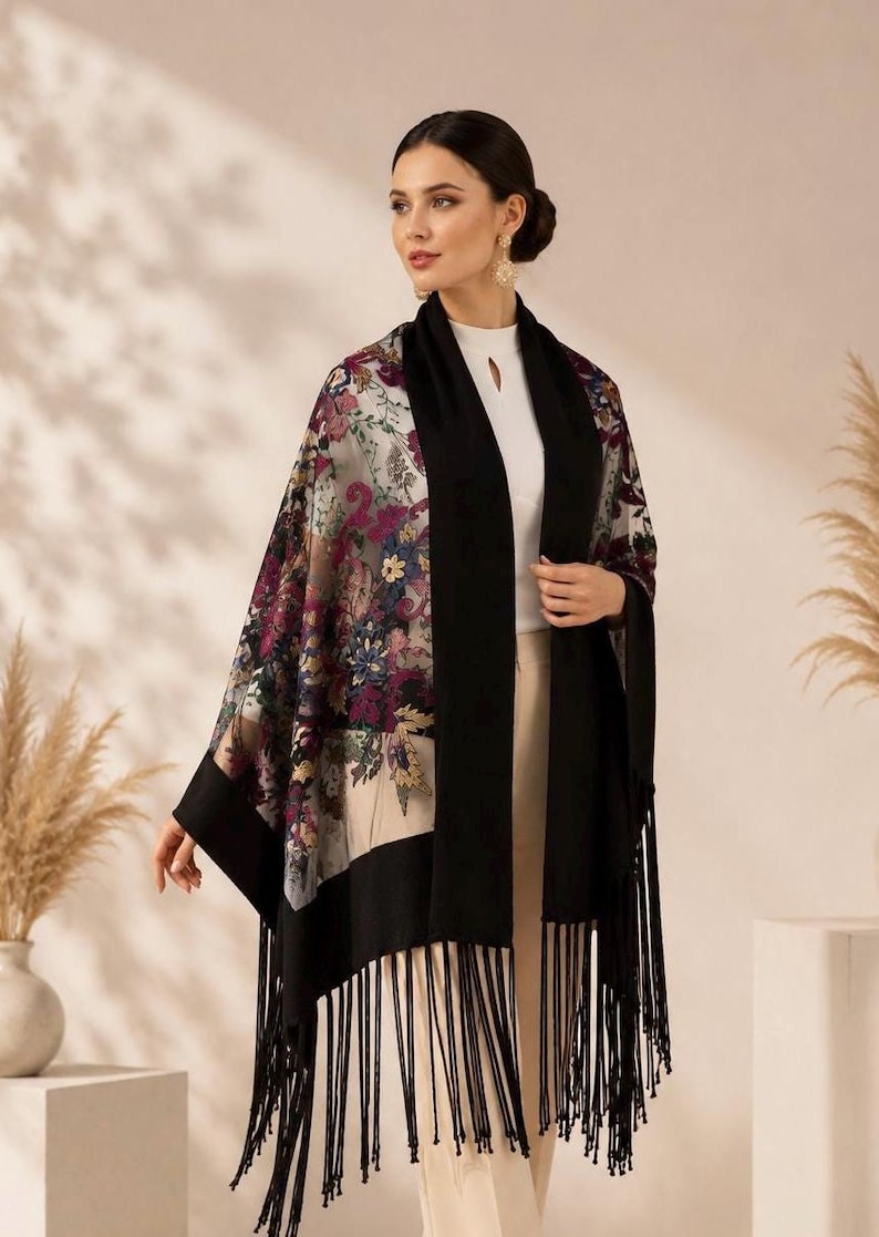 A floral embroidery shawl with black borders and fringe. The shawl features a colorful floral pattern on a light background. The black borders frame the floral design, and the fringe adds a decorative element. This accessory can be worn over the shoulders.