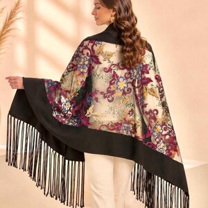A sheer shawl with a black border and fringe. The shawl features a floral pattern in shades of purple, blue, and gold. The model is wearing the shawl over a black top and white pants.