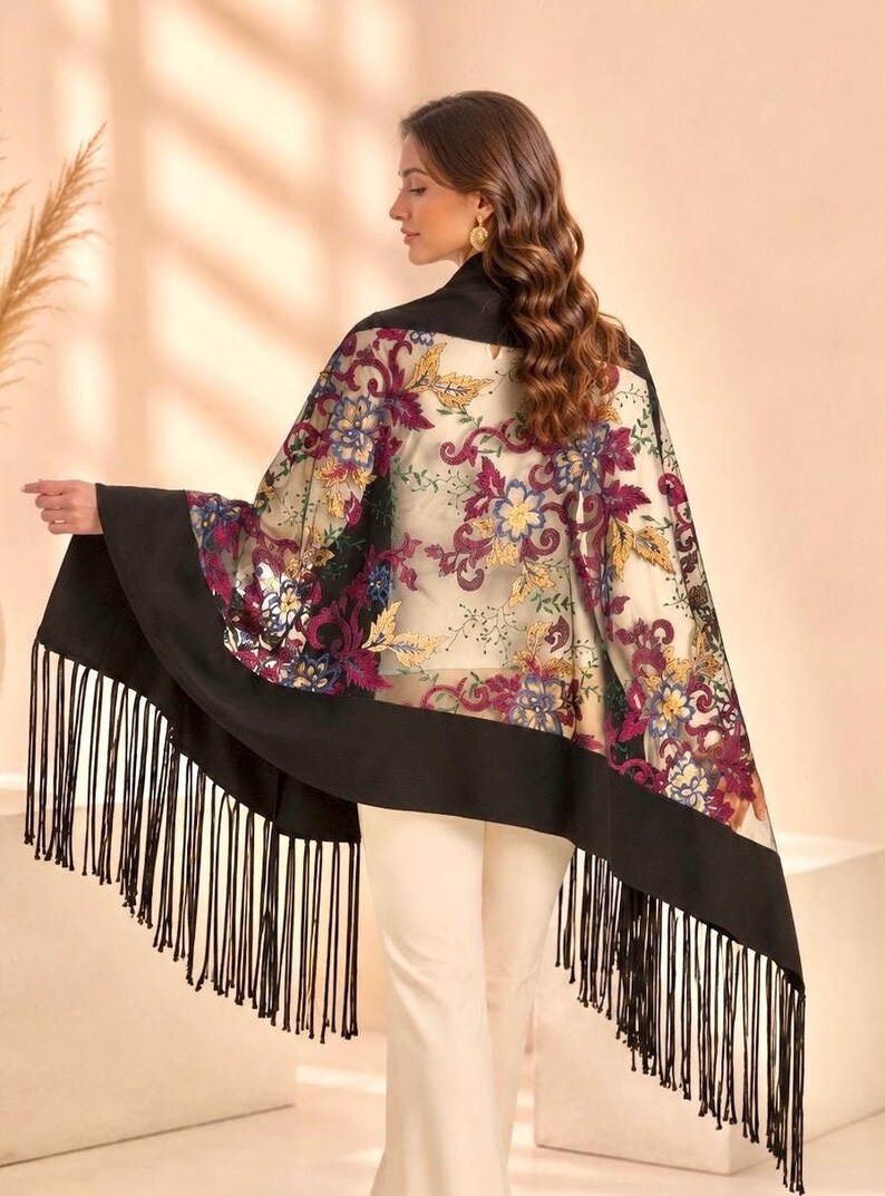 A sheer shawl with a black border and fringe. The shawl features a floral pattern in shades of purple, blue, and gold. The model is wearing the shawl over a black top and white pants.