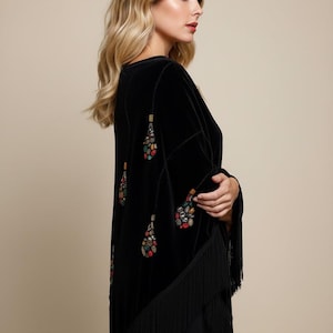 Black velvet shawl with colorful beaded embellishments and fringe trim. The long sleeves and open front create a flowing silhouette. A stylish and elegant garment.