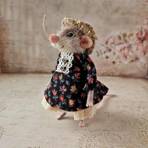 Puede incluir: Una figura de ratón de fieltro hecha a mano, vestida con un vestido negro con estampado floral y un cuello de encaje blanco. El ratón tiene un cuerpo marrón claro, una nariz rosa y un peinado rizado de color marrón claro. El vestido tiene un dobladillo con volantes.