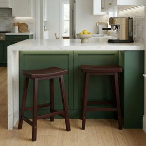 May include: Two dark brown wooden bar stools with curved seats are positioned in front of a green kitchen island. The island has a white countertop and a built-in coffee maker. The stools are on a light wood floor.